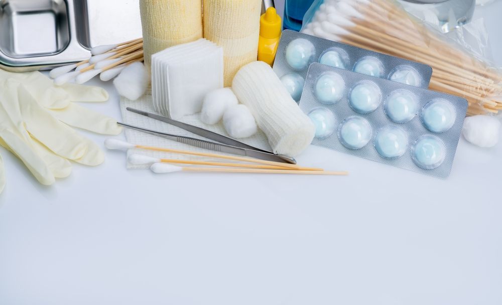Medical supplies on a white surface, including sterile gloves, gauze, cotton balls, tweezers, and blister-packed pills.
