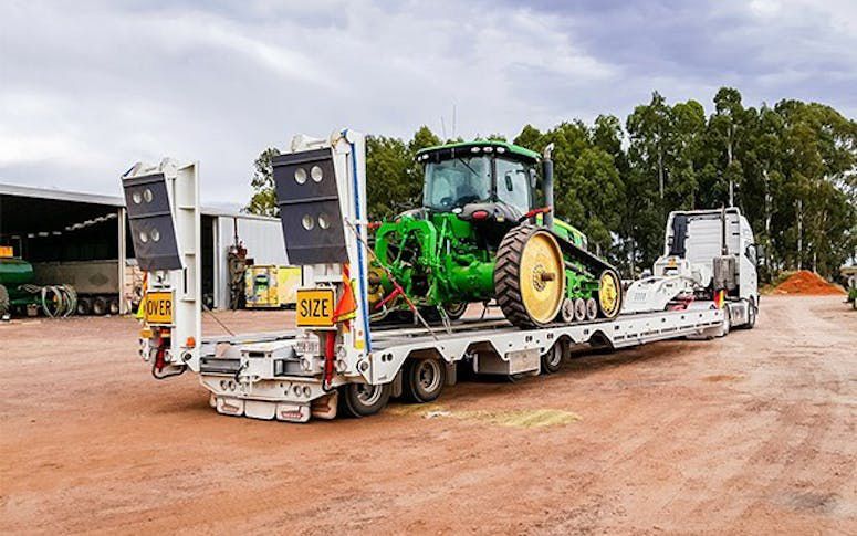 Green tractor on a flatbed trailer, being transported on a dirt road.