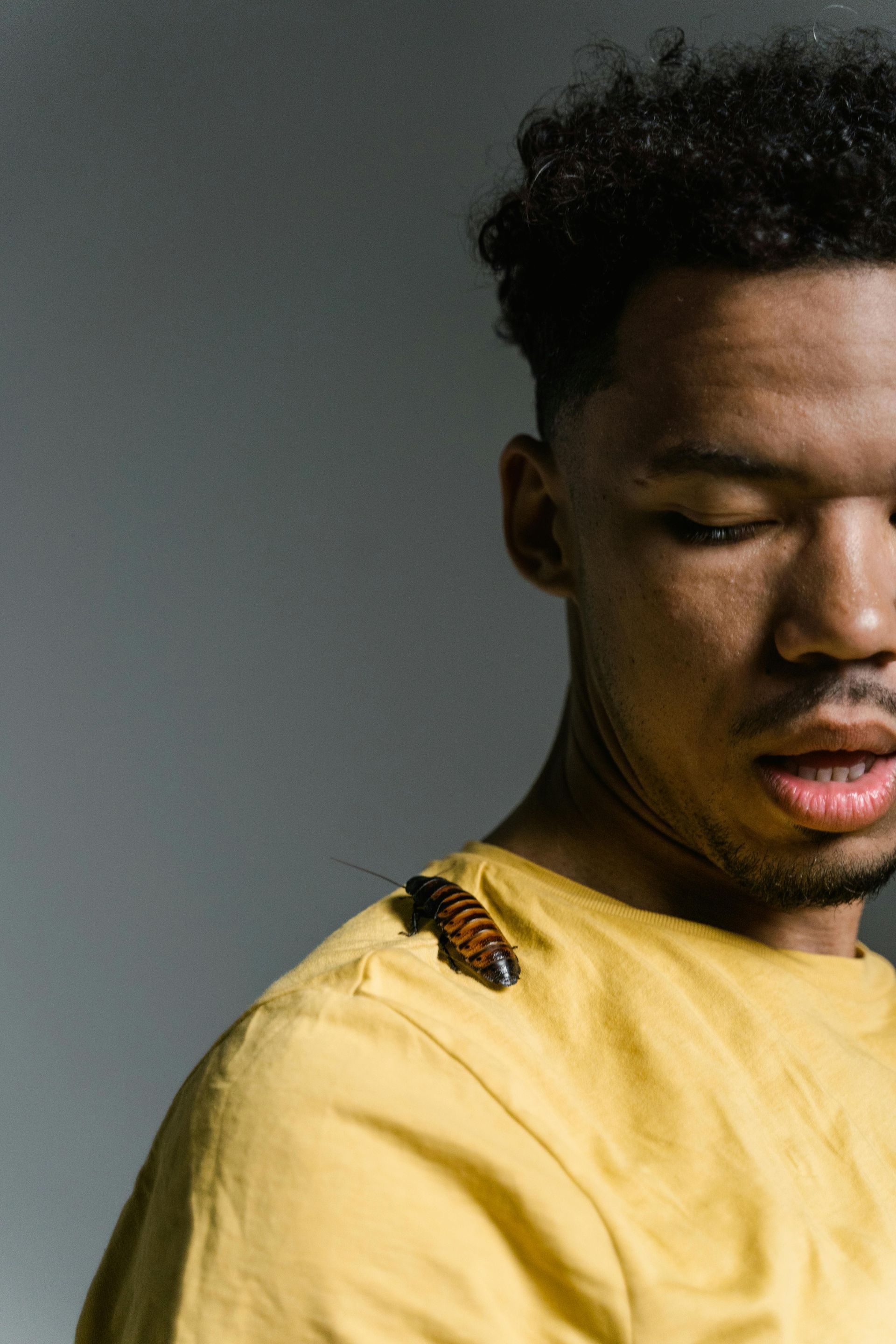 Man with a shocked expression, wearing a yellow shirt, has a cockroach on his shoulder. Gray background.