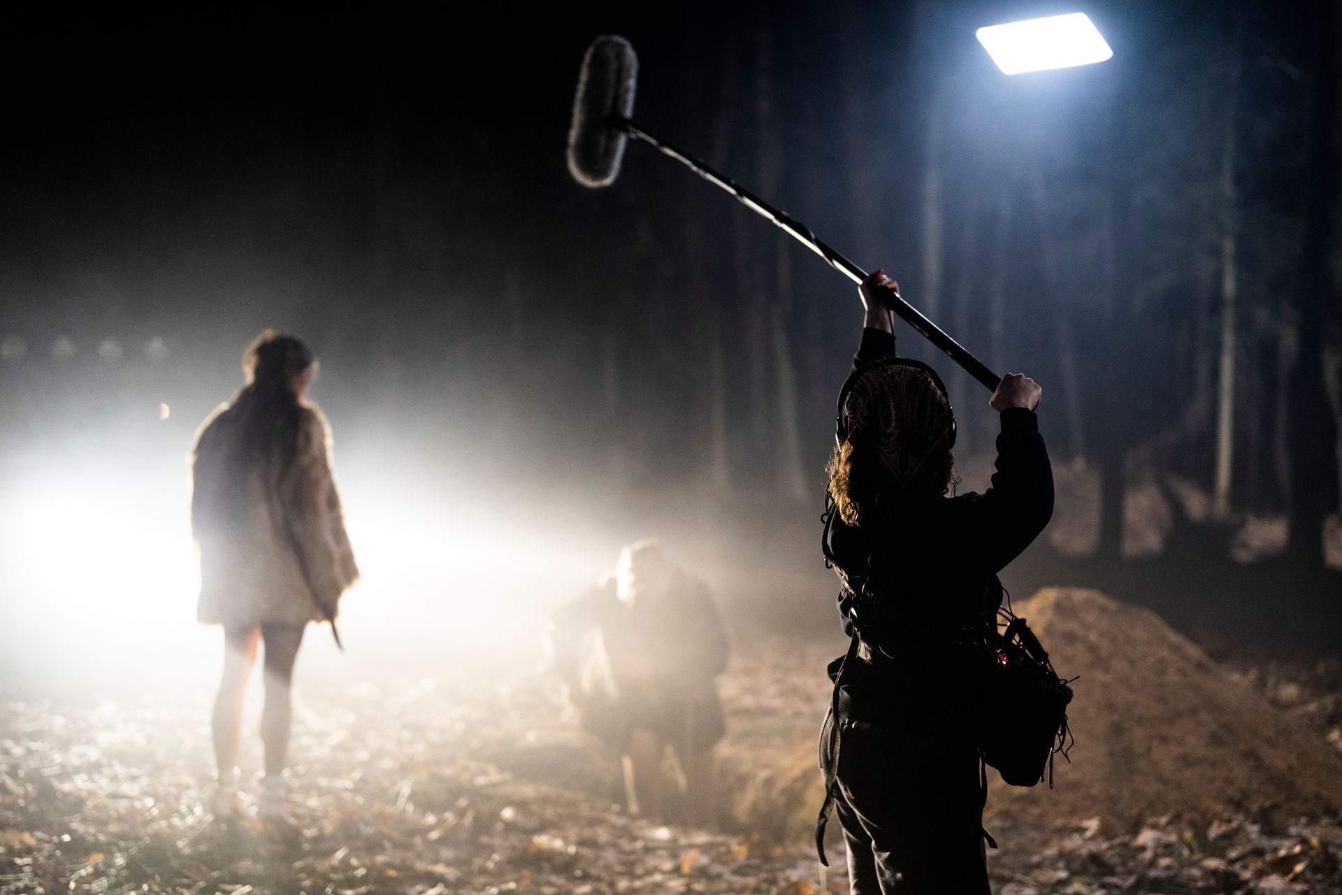 Een geluidstechnicus houdt een microfoon gericht op acteurs in een mistige nachtelijke buitenscène bij een grote schijnwerper