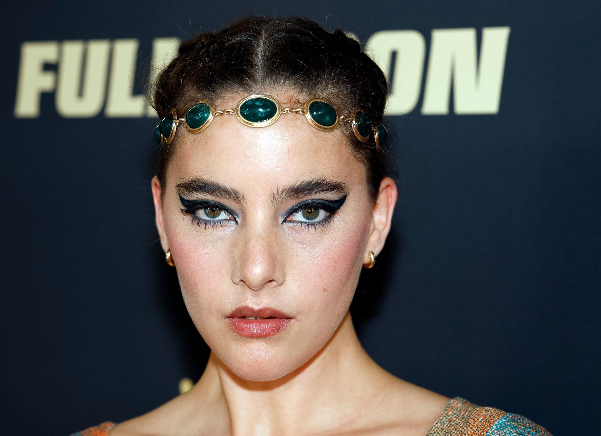 Vrouw met donker haar, een met juwelen versierde haarband en opvallende oogmake-up, kijkt in de camera.