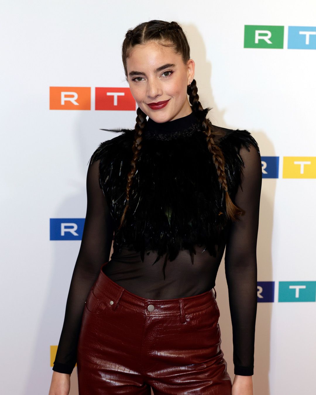 Vrouw met gevlochten haar, gekleed in een zwarte top en bordeauxrode broek, lachend poseert voor witte achtergrond met logos.