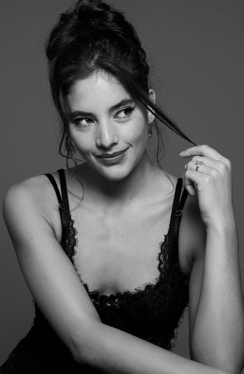 Vrouw in een kanten topje, opgestoken haar, lachend en spelend met een plukje haar, tegen een neutrale achtergrond.