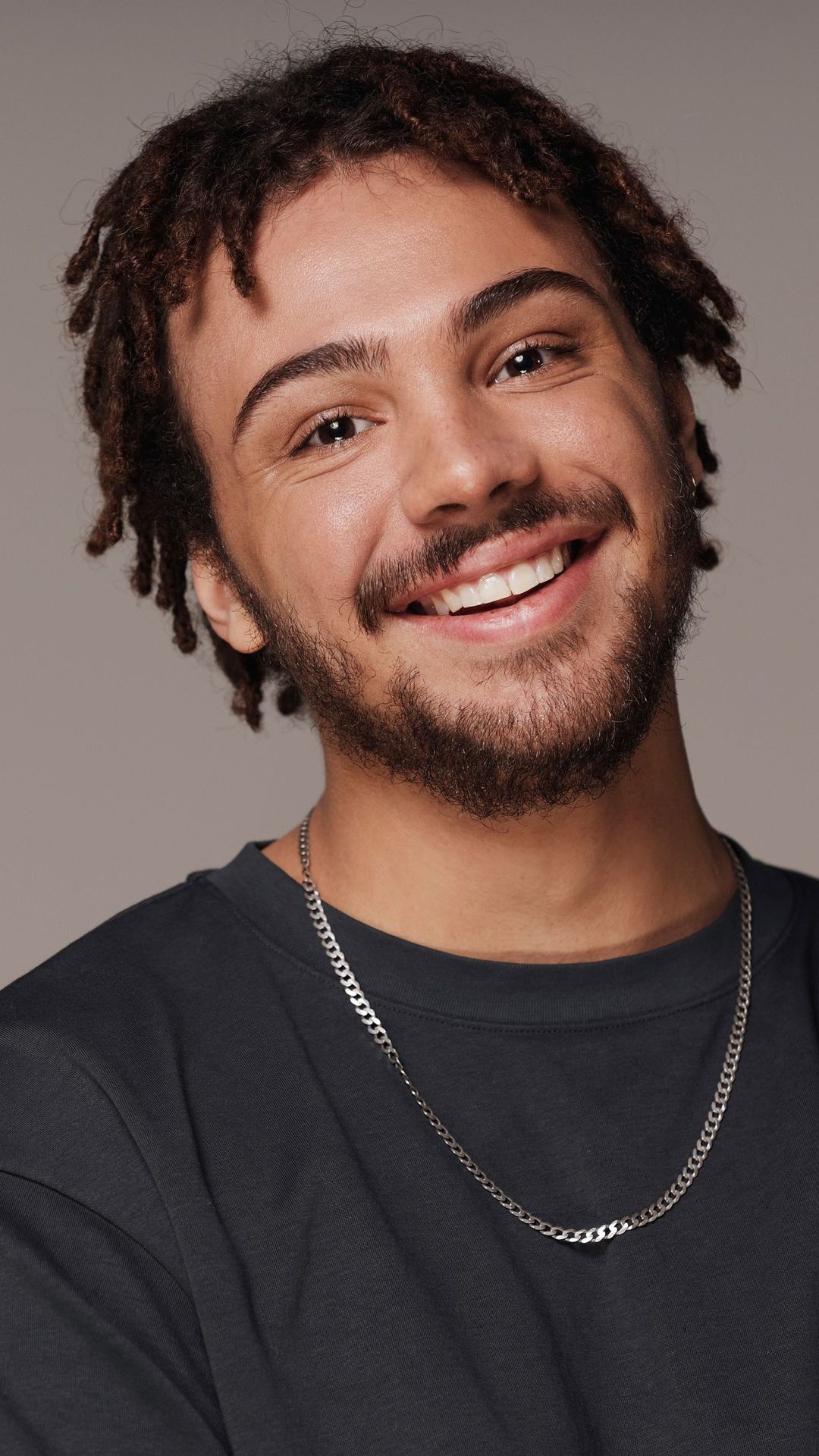 Een lachend persoon met bruine dreadlocks en een baard, gekleed in een zwart shirt en met een zilveren ketting.