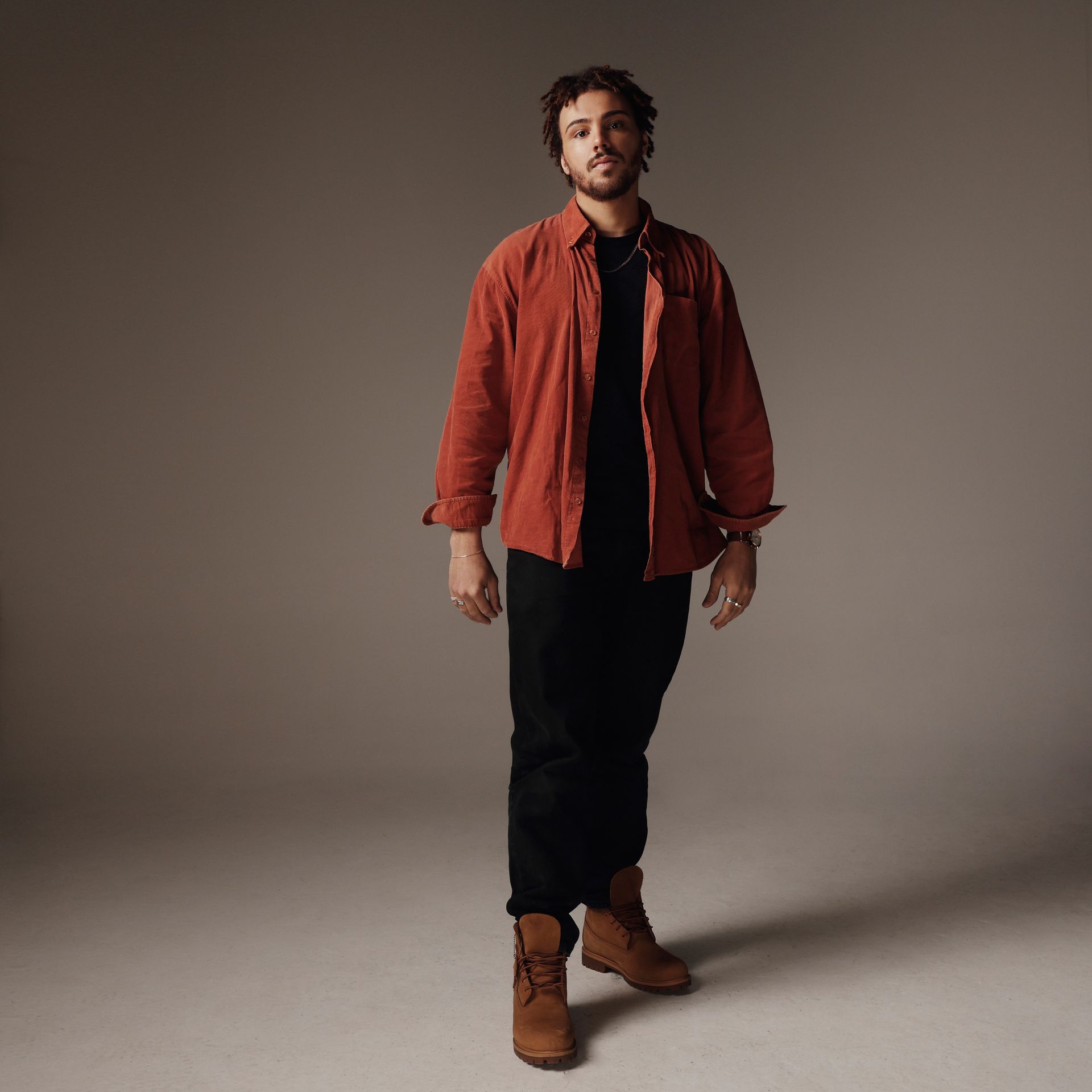 Een man in een oranje shirt, zwarte broek en bruine laarzen kijkt in de camera tegen een neutrale achtergrond.