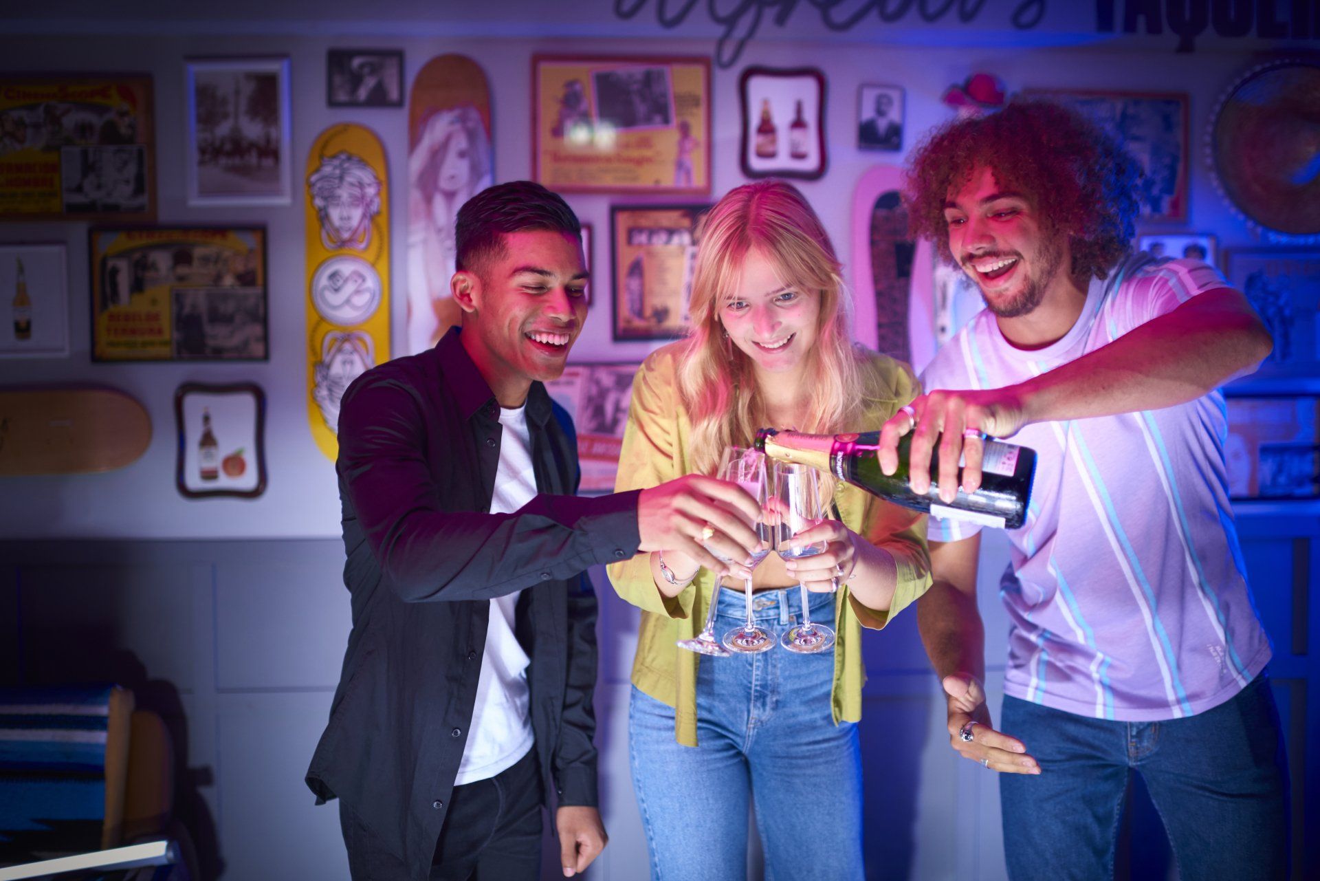 Drie mensen proosten met champagne in een kamer versierd met skateboards, terwijl ze lachend een fles leegschenken.