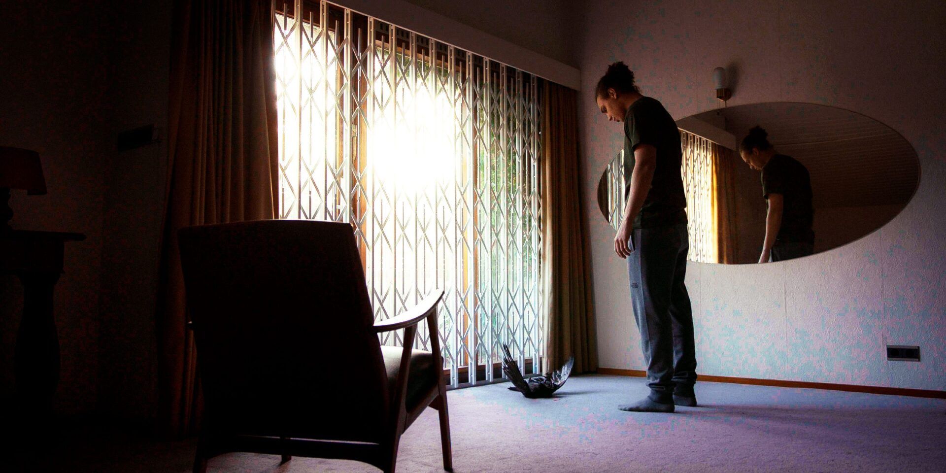 Een persoon staat in een kamer en kijkt naar de vloer; op de achtergrond zijn een ovale spiegel en gordijnen te zien.