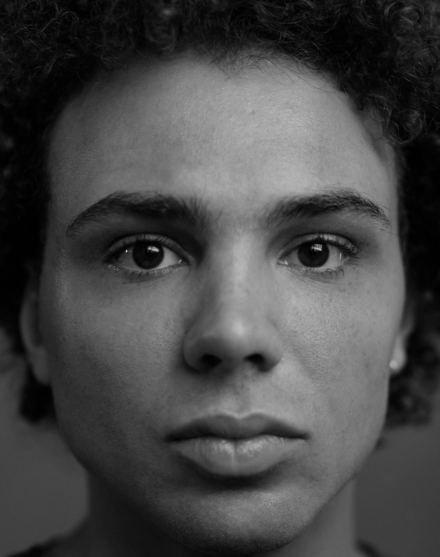 Close-up zwart-witportret van een persoon met krullend haar, die de kijker recht aankijkt.