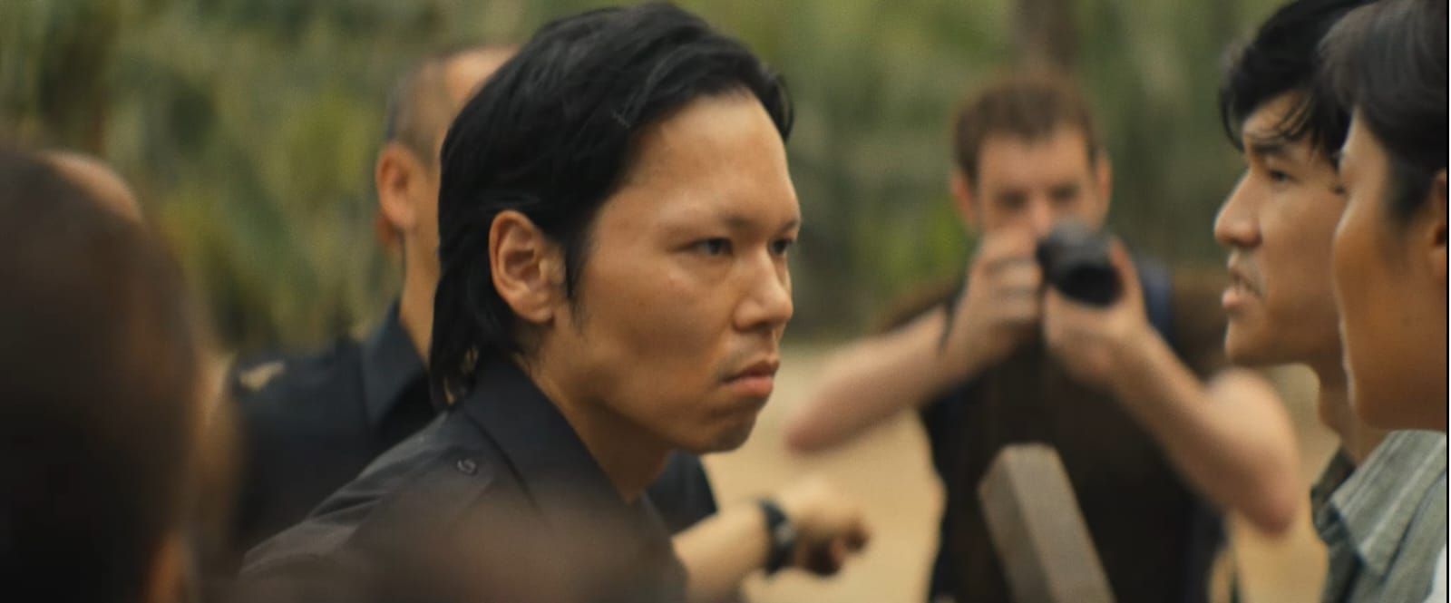 Een man in een zwart shirt kijkt naar rechts, omringd door andere mensen in de buitenlucht.