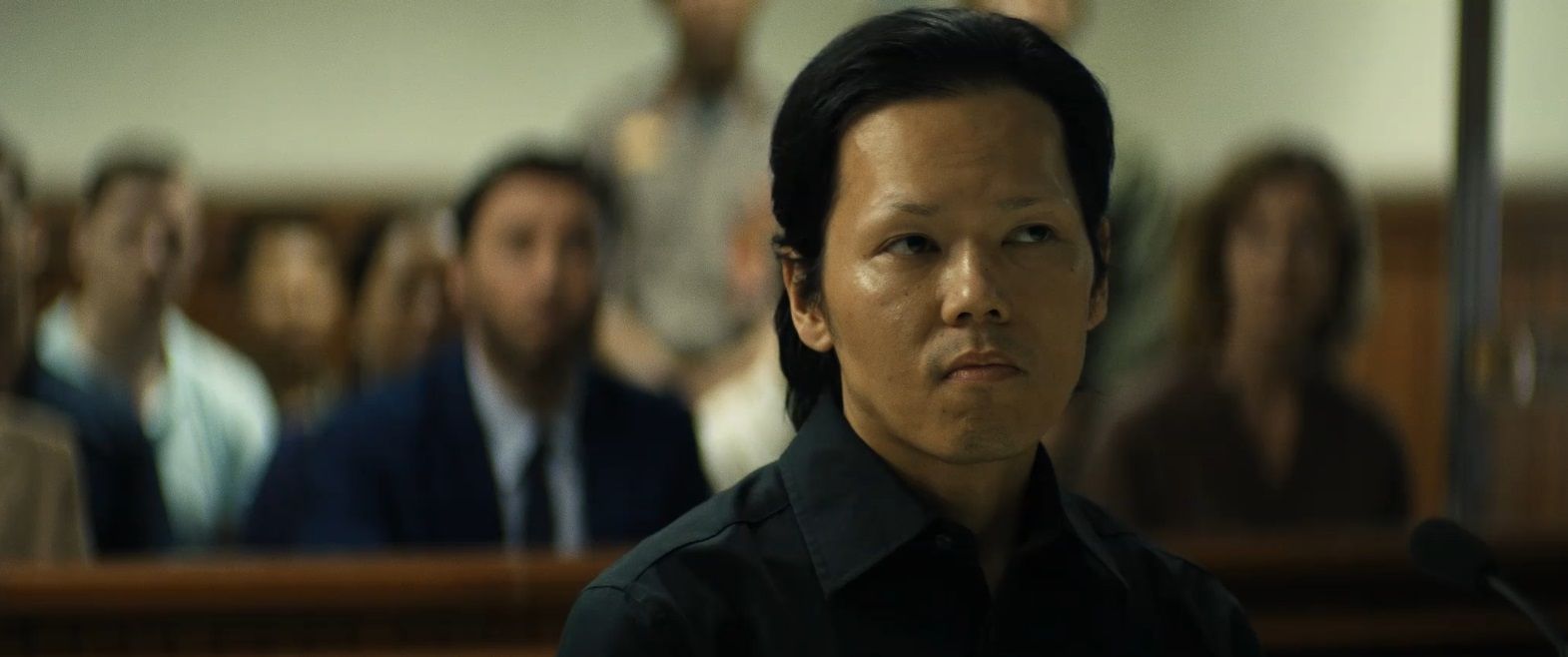 Een man in een zwart shirt zit in een rechtszaal en kijkt opzij. Op de achtergrond zijn andere mensen wazig te zien.