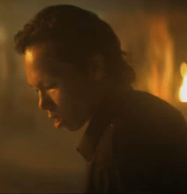 Een man met donker haar kijkt naar beneden in een schemerige, vurige omgeving.