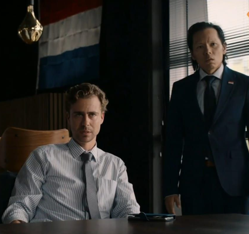 Twee mannen in een kantooromgeving. De ene zit met een serieuze blik, de andere staat. Een vlag hangt aan de muur.