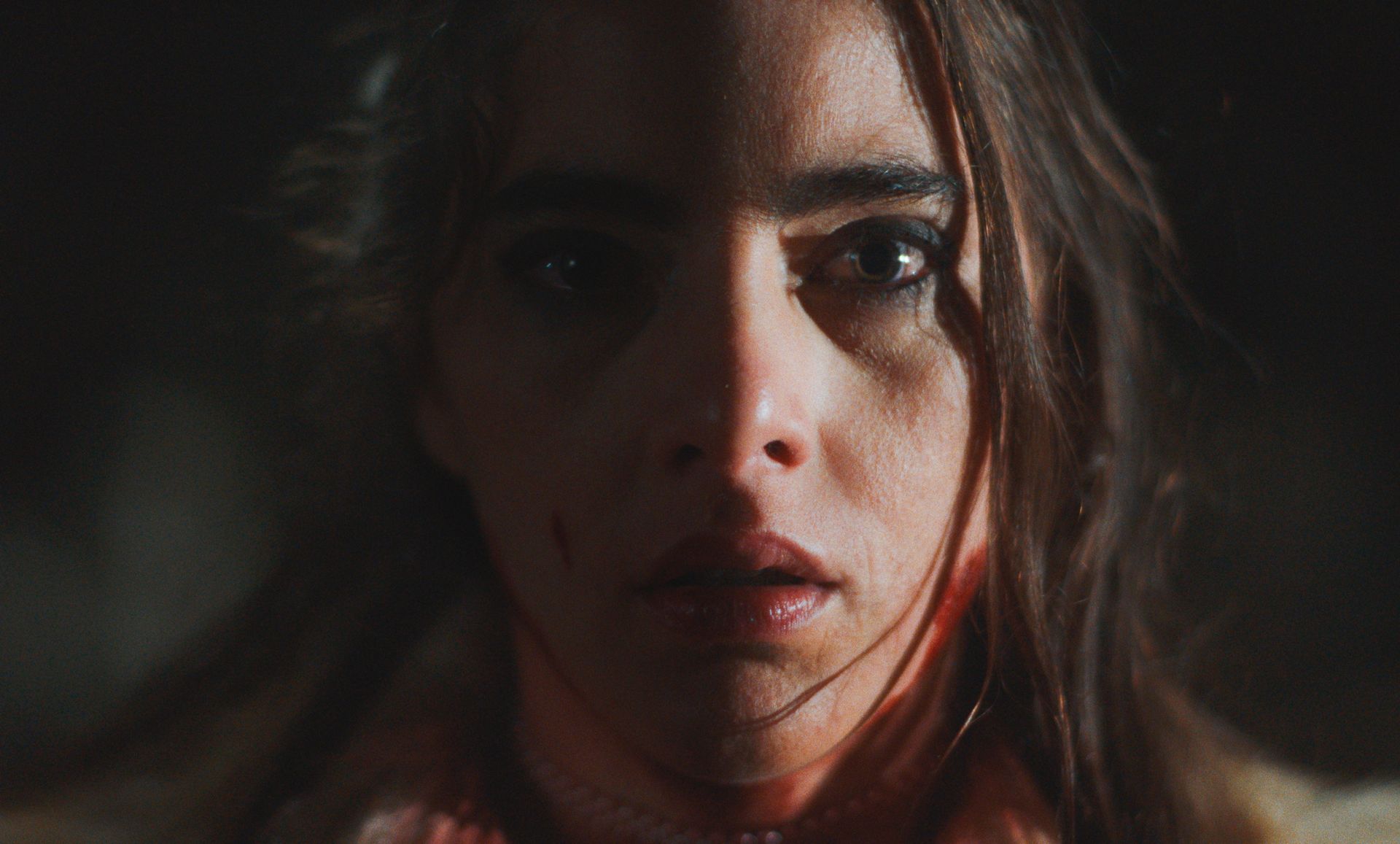 Vrouw met een bebloed gezicht, donker haar en grote ogen, in een schemerig verlicht interieur.