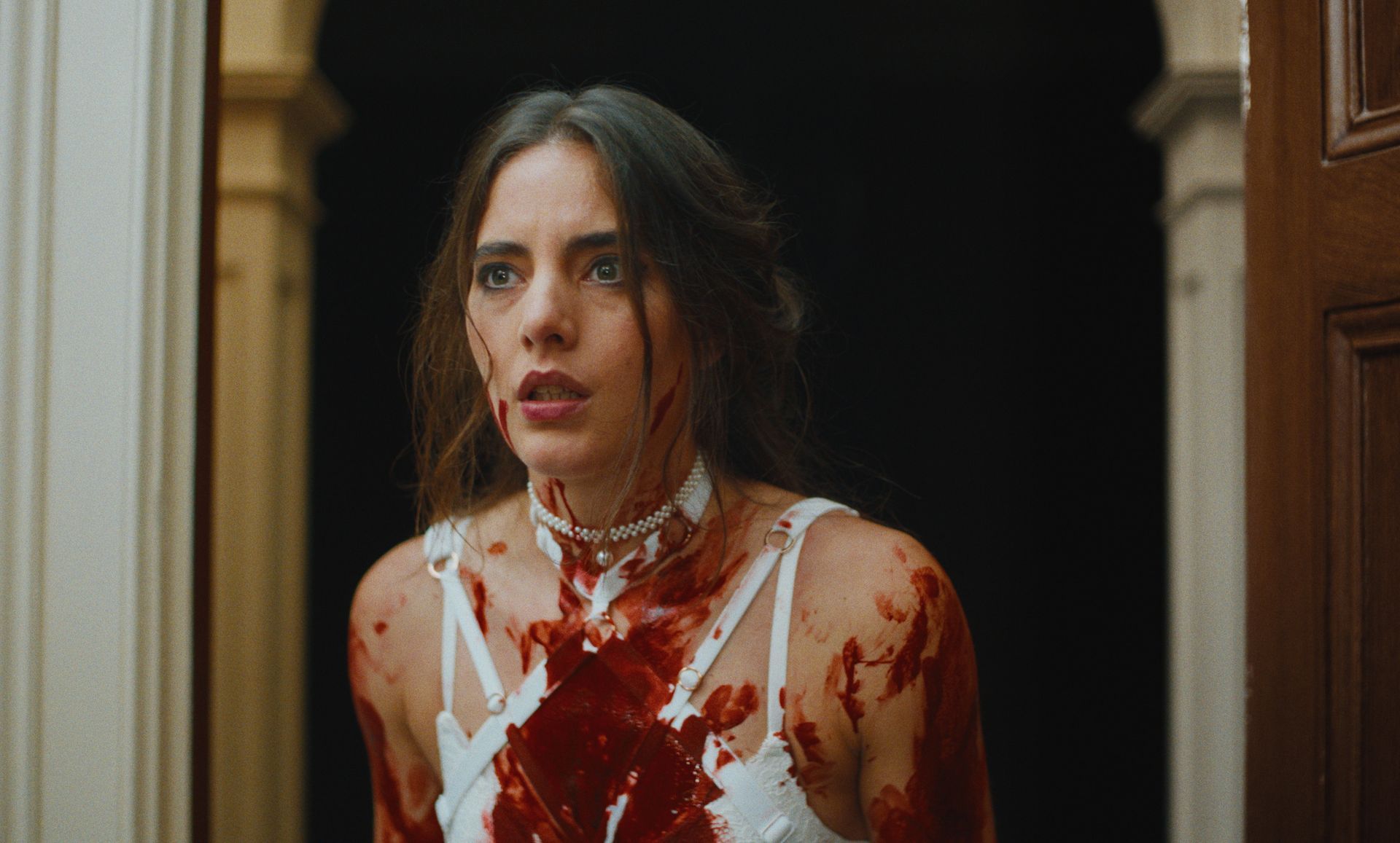 Een vrouw in witte lingerie, onder het bloed, staart met grote ogen vanuit een deuropening.