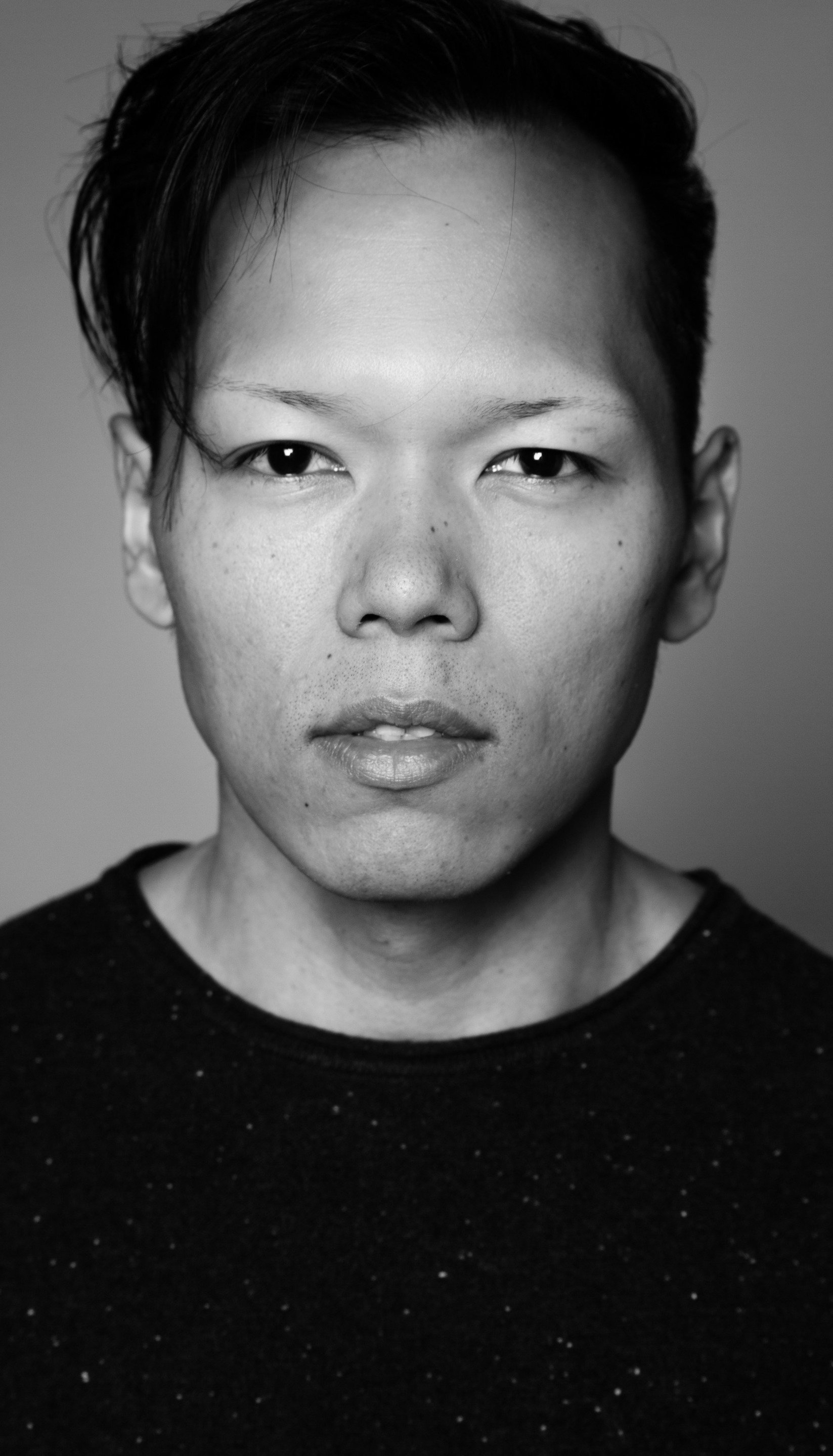 Een man met achterovergekamd zwart haar, gekleed in een donkere trui, kijkt recht vooruit op een zwart-witfoto.