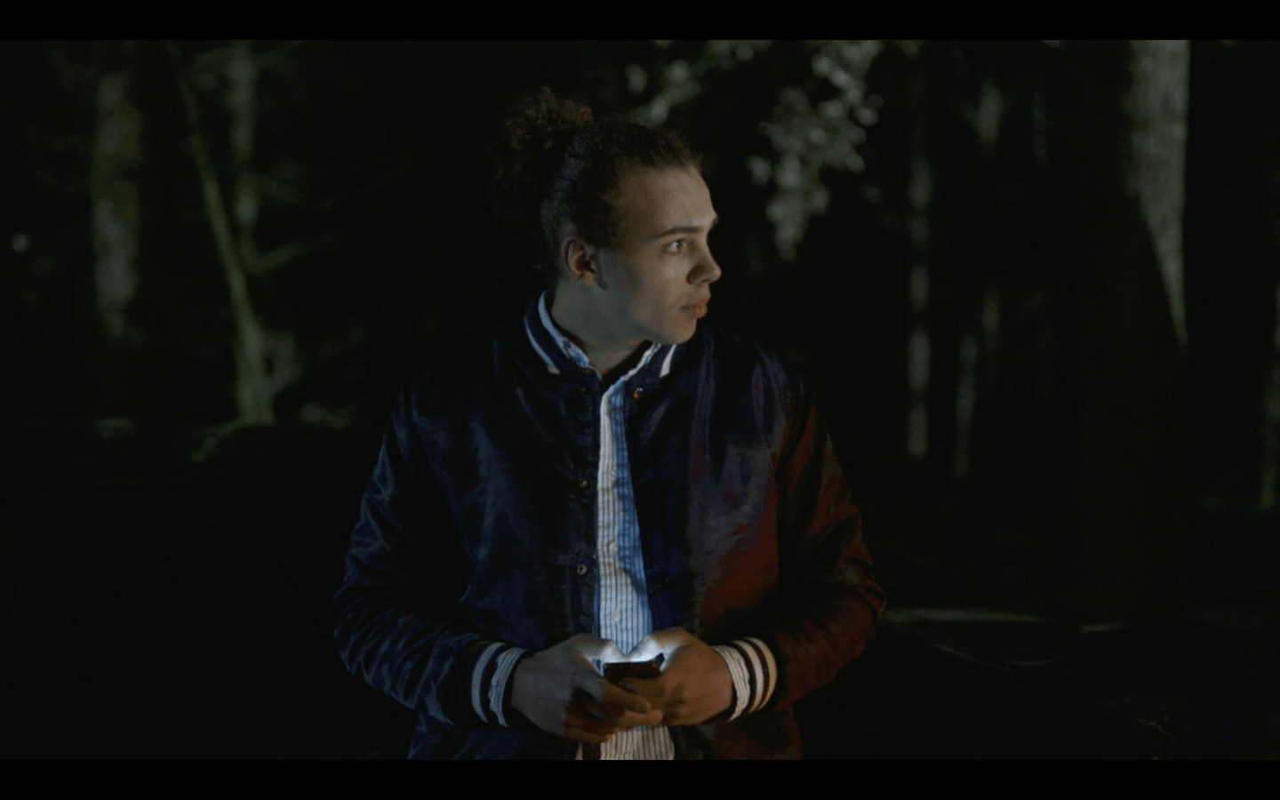 Een man in een donker bos, met een telefoon in zijn hand, kijkt bezorgd opzij.