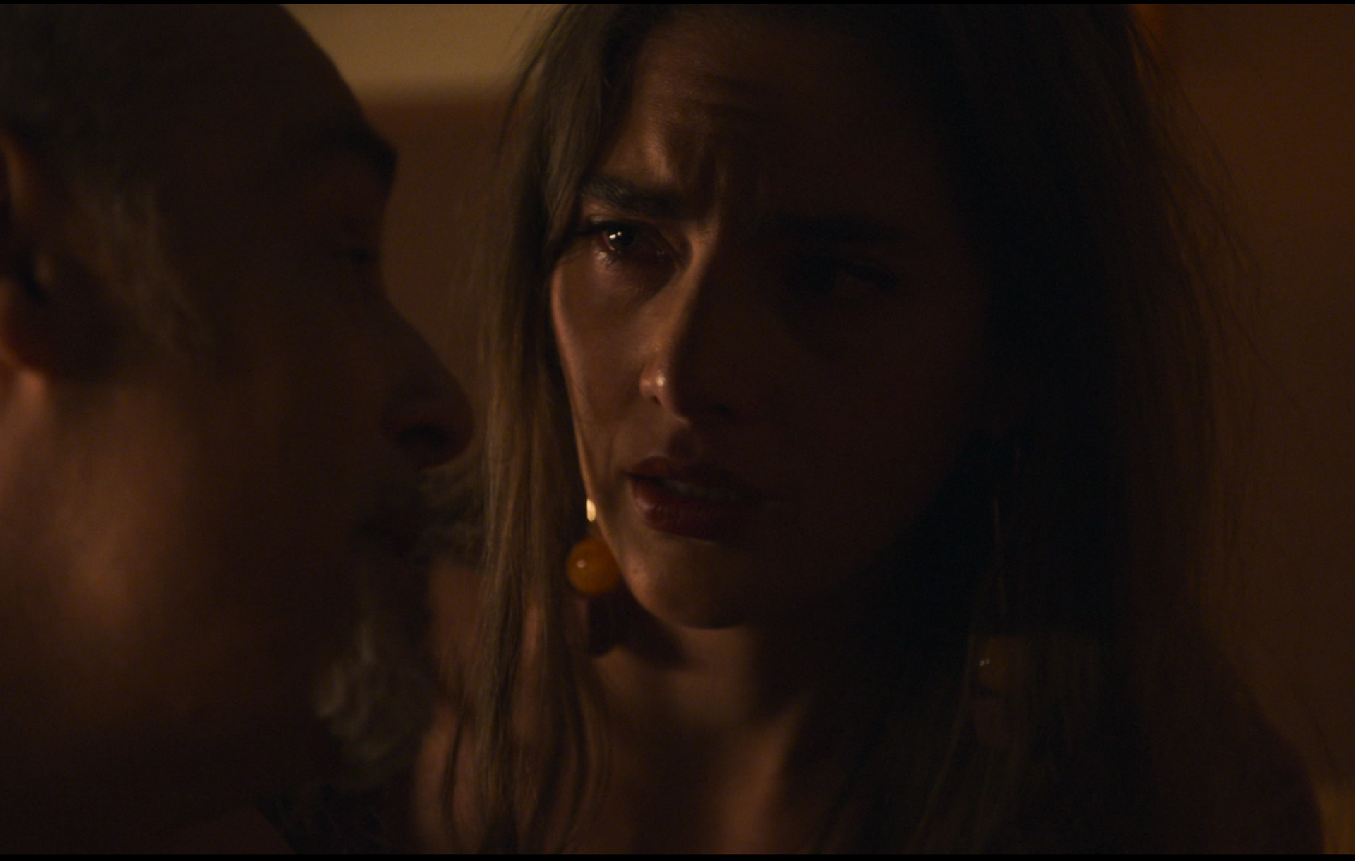 Een vrouw met lang haar en grote oorbellen kijkt bezorgd en praat met iemand buiten beeld in een schemerig verlichte ruimte.