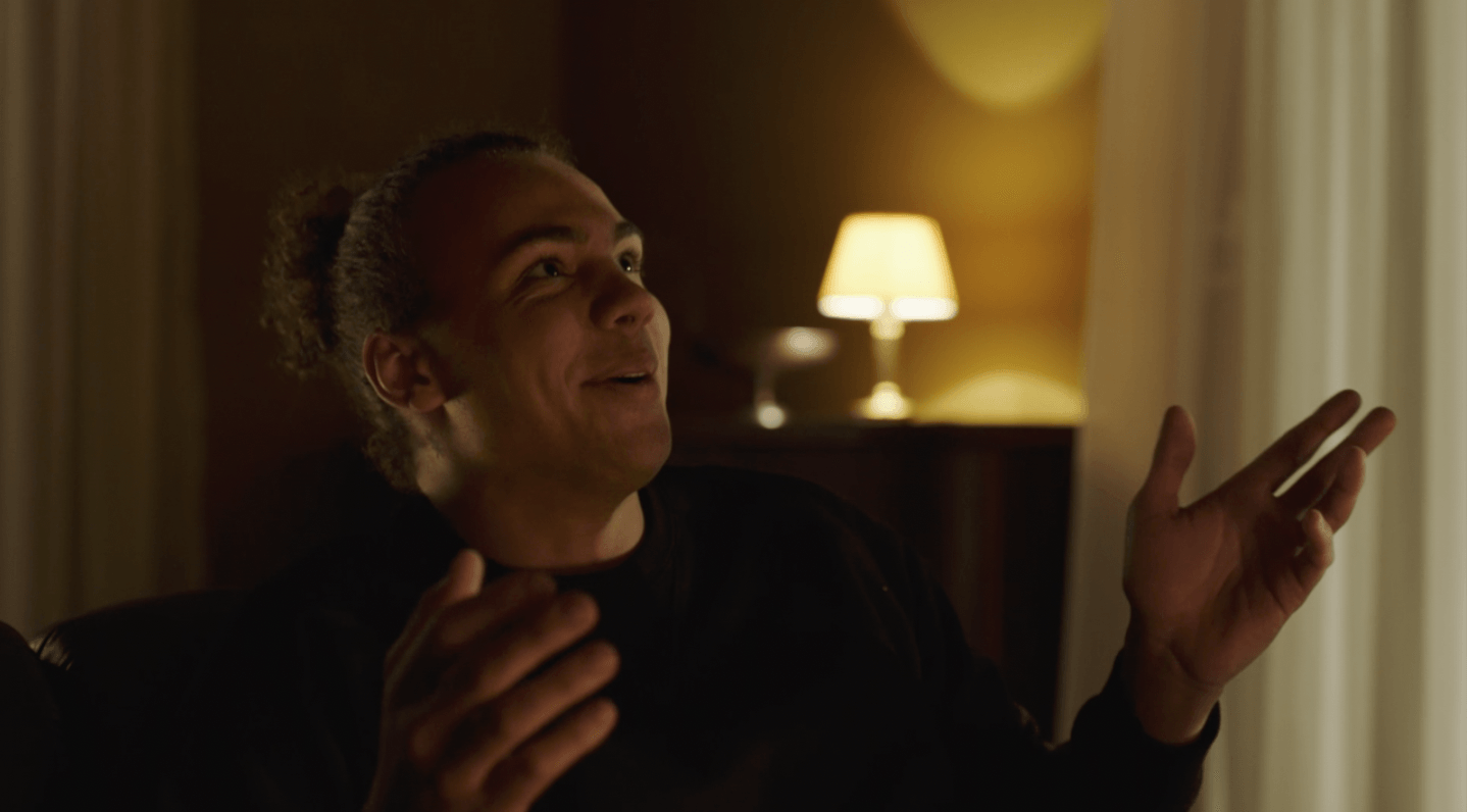 Een man met een paardenstaart gebaart en lacht, zittend in een kamer vlakbij een lamp.