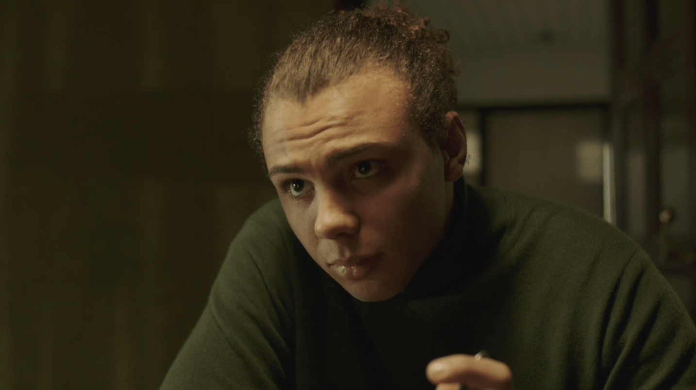 Man met knotje, gekleed in een groene coltrui, kijkt aandachtig. Donkere achtergrond, mogelijk binnen.