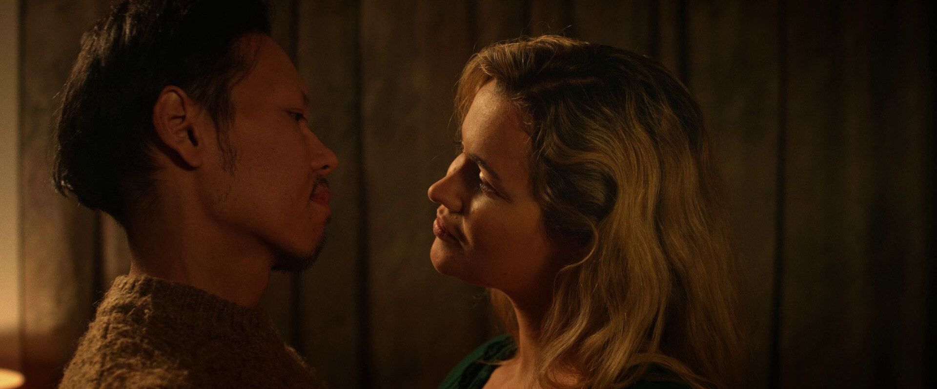 Twee mensen staan dicht tegenover elkaar, op het punt elkaar te kussen in een schemerige omgeving.