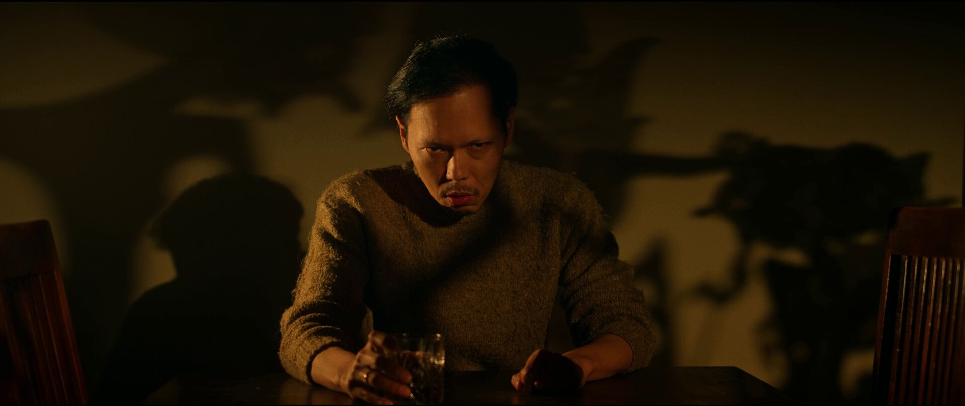 Man zit aan een tafel en houdt een glas vast. Schaduw op de achtergrond. Warm licht.