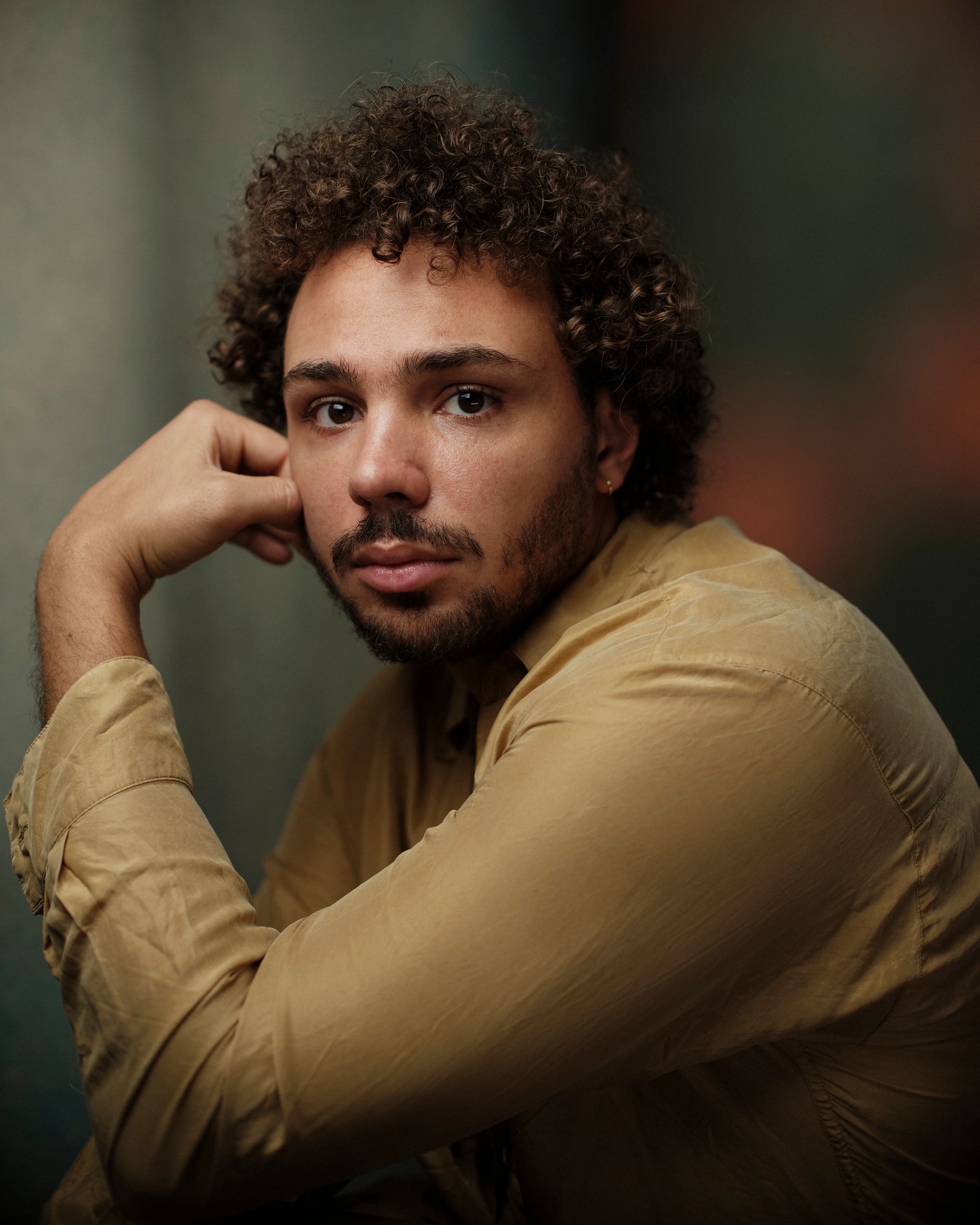 Man met krullend haar, hand voor zijn gezicht, gekleed in een beige shirt, kijkt in de camera.