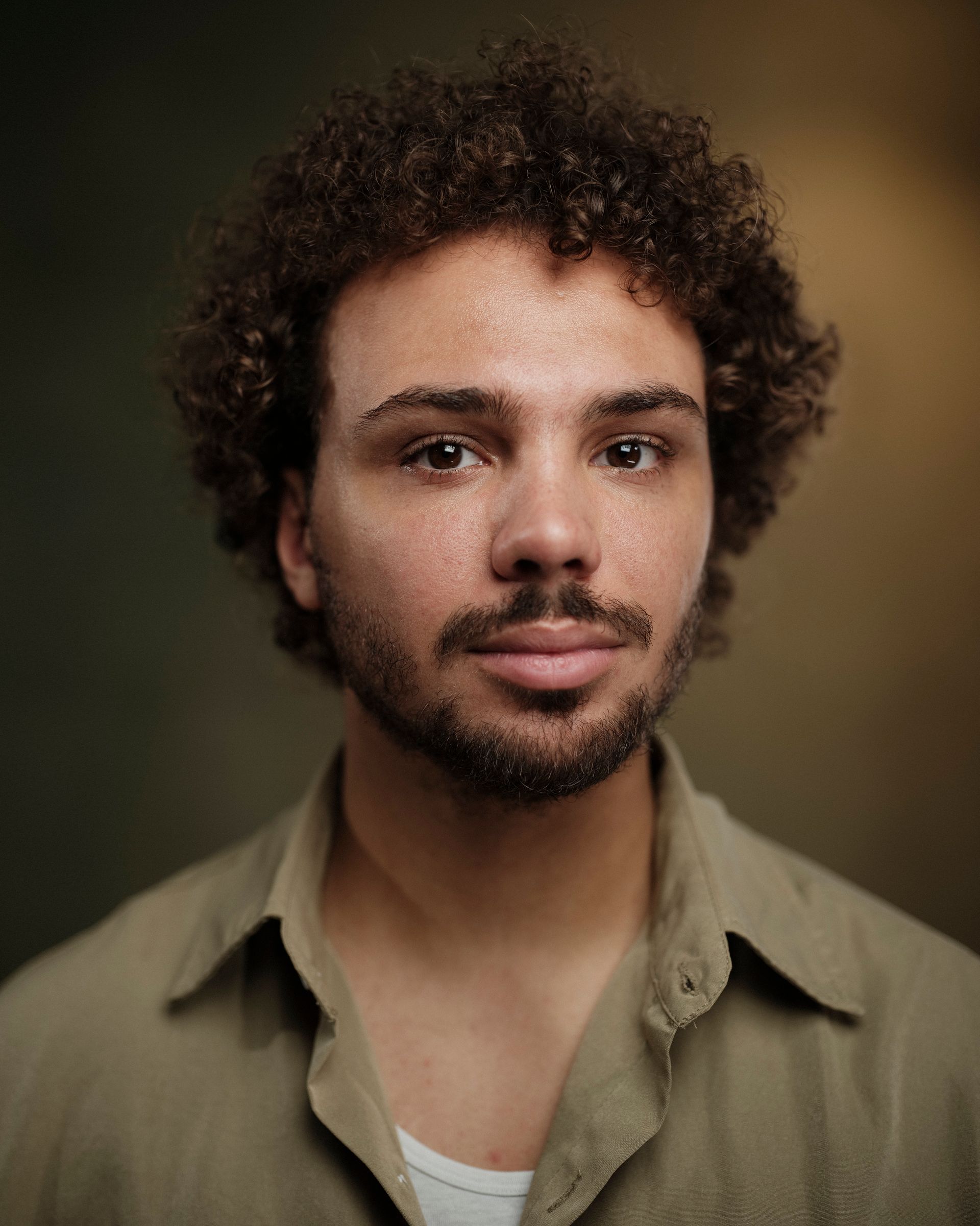 Man met krullend haar en een baard, gekleed in een beige shirt, kijkt in de camera. Donkere, wazige achtergrond.