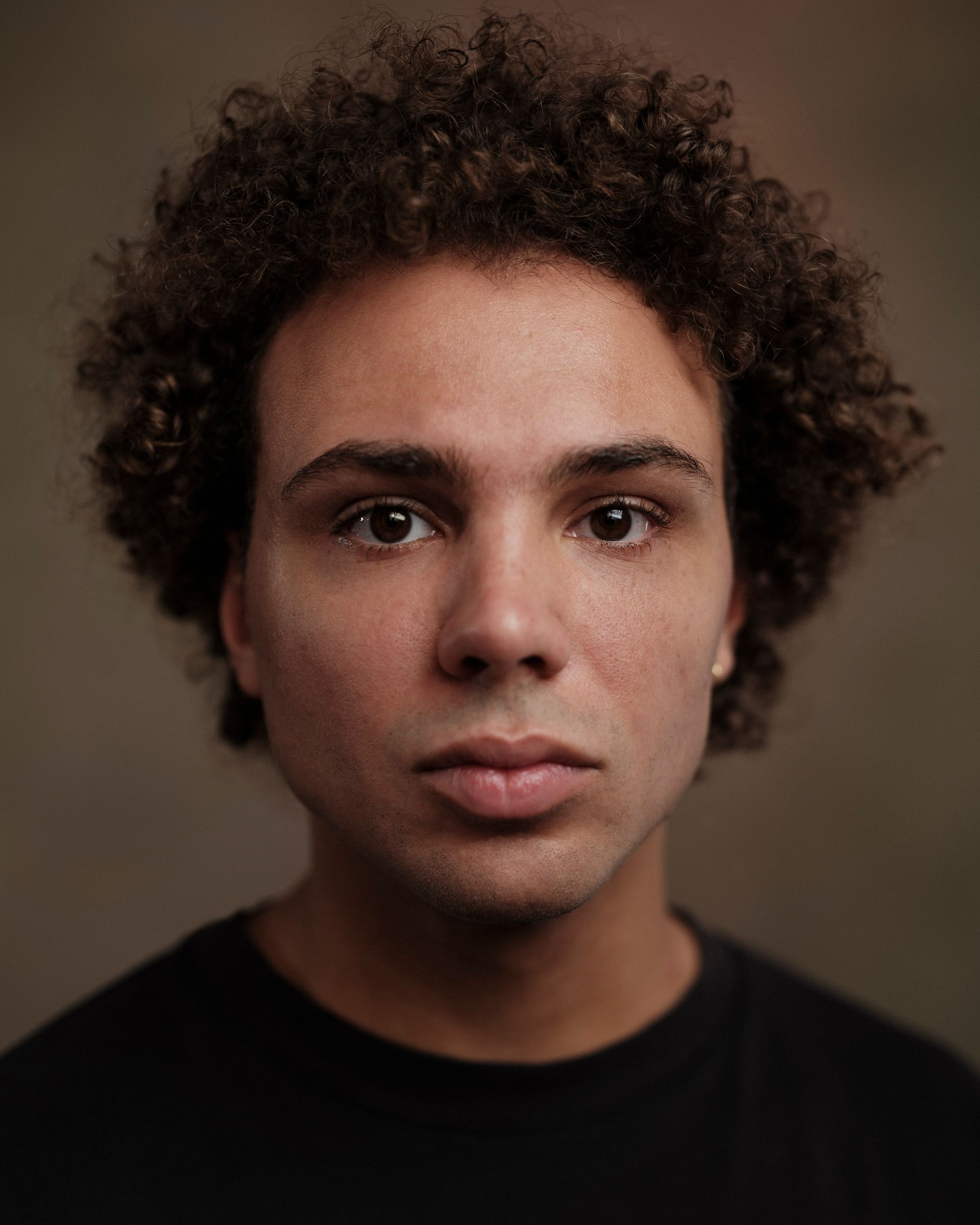 Persoon met krullend bruin haar, die de kijker recht aankijkt en een zwart shirt draagt.