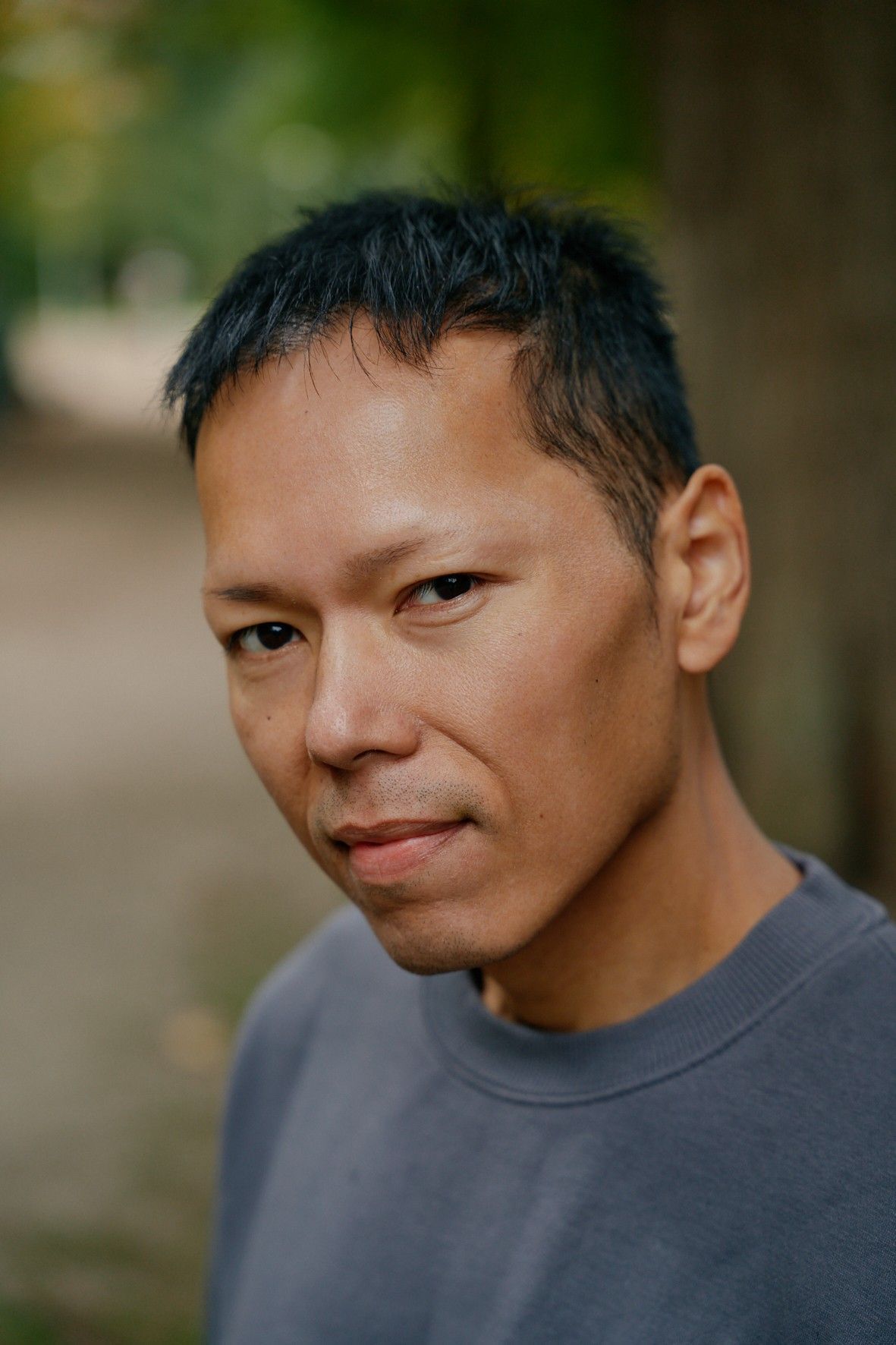 Man met donker haar en een grijs shirt kijkt naar de camera; wazige buitenachtergrond.