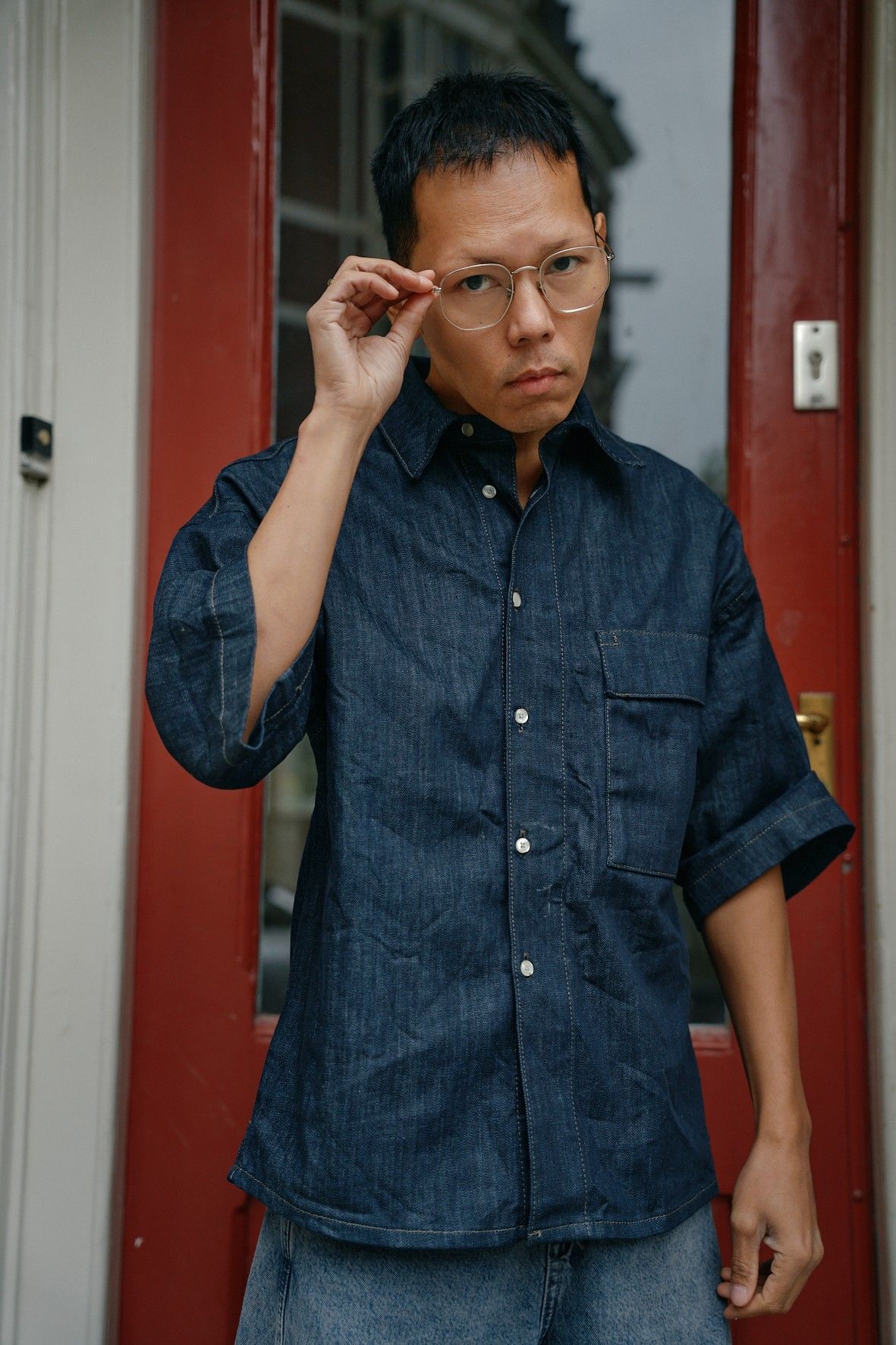 Een man met een bril en een spijkerhemd aan, die zijn bril rechtzet bij een rode deur.