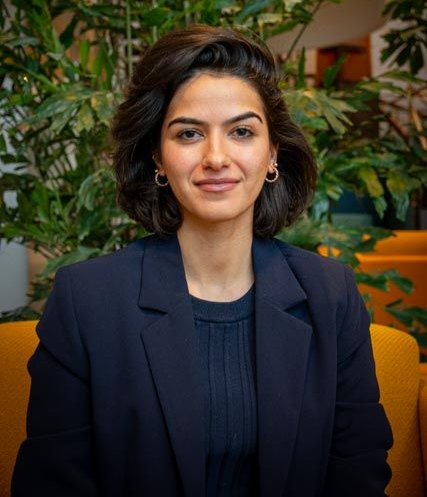 Vrouw met kort donker haar, gekleed in een donkerblauwe blazer, lacht voor een groene plant.