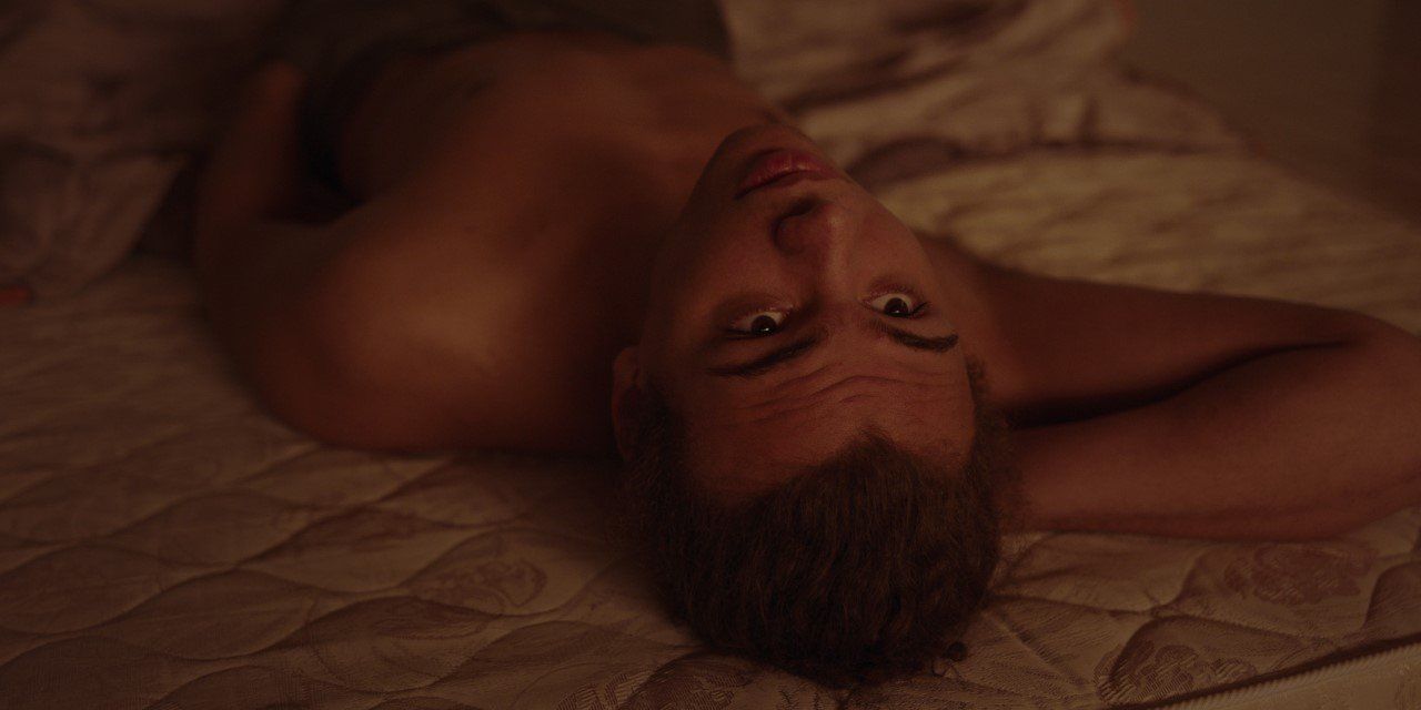 Een persoon met ontbloot bovenlichaam ligt op een bed en kijkt met een peinzende uitdrukking naar boven.
