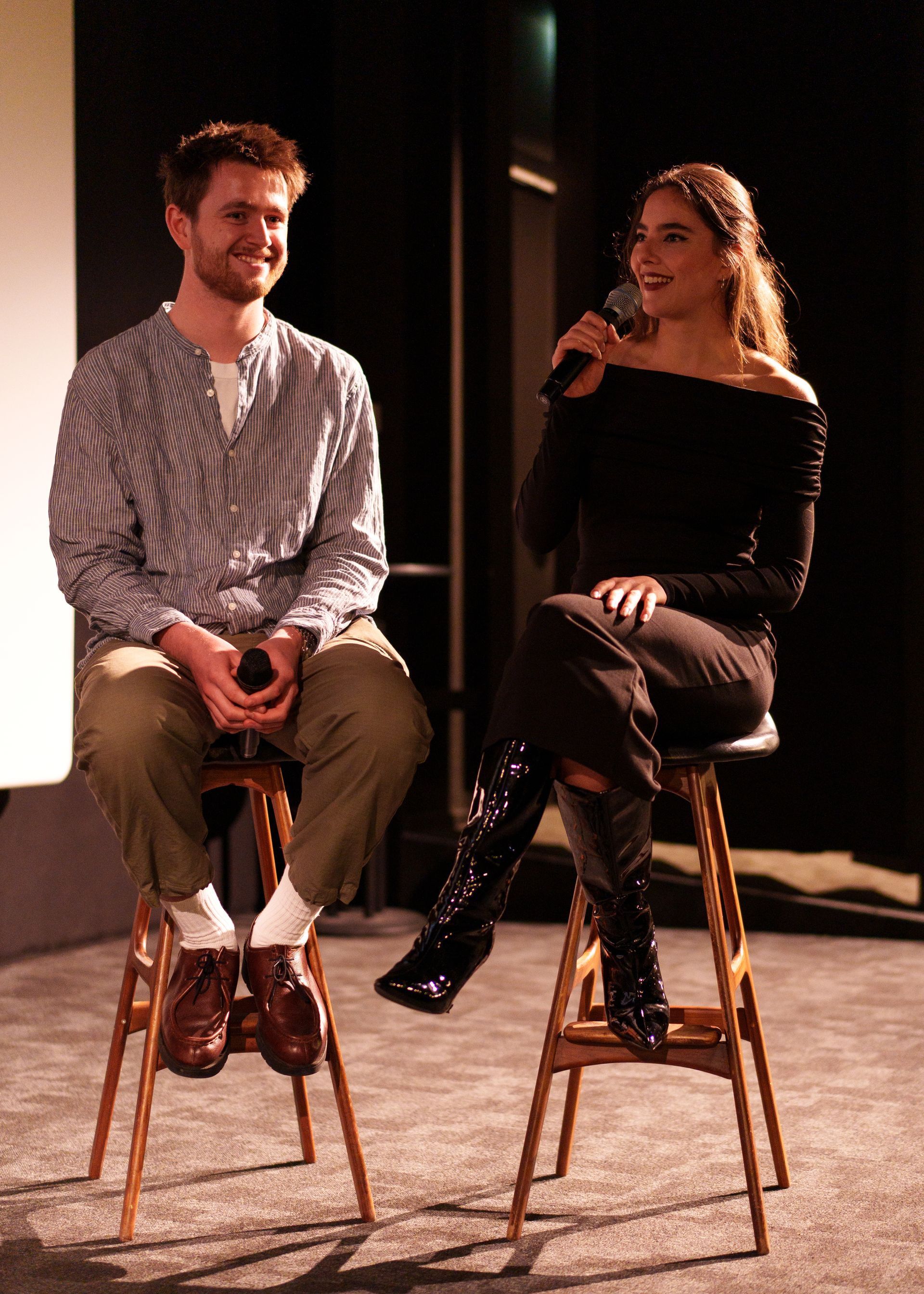 Twee mensen zitten op krukjes en voeren een gesprek. De een houdt een microfoon vast, de ander glimlacht. Donker interieur.