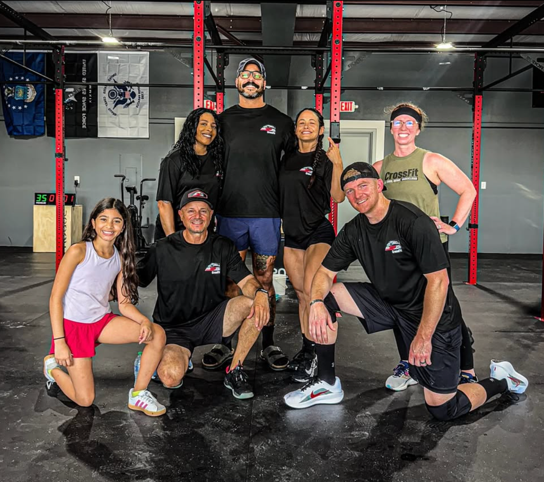 Group of people posing in a gym. Some are kneeling, wearing athletic gear. A child is kneeling.