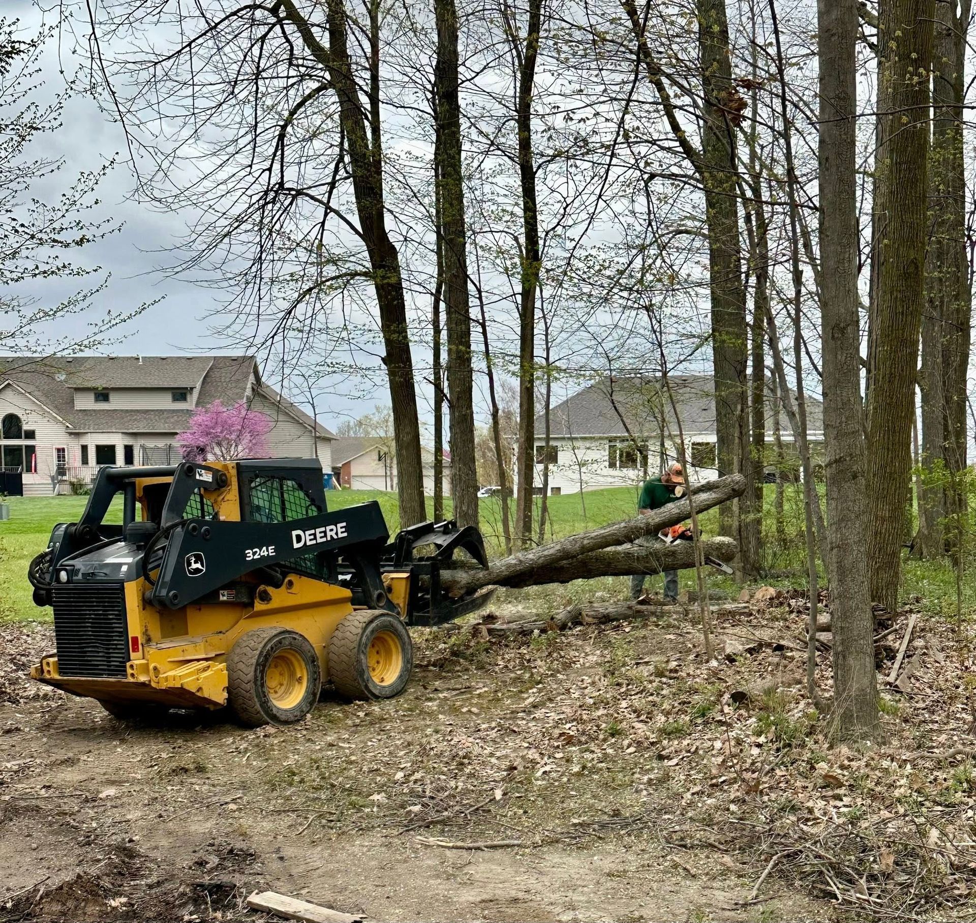 A man is standing next to a skid steer in the woods.