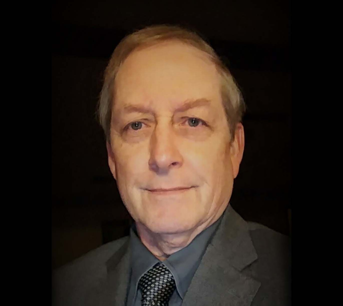 Man in a gray suit and tie, smiling slightly, against a dark background.