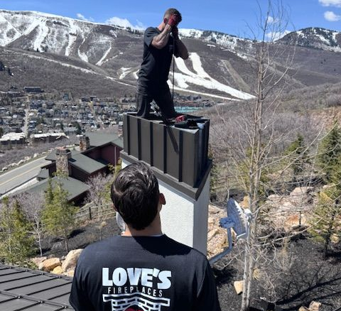 Two chimney sweeps on a roof, working on a brick chimney. One is reaching, the other is holding a hose.
