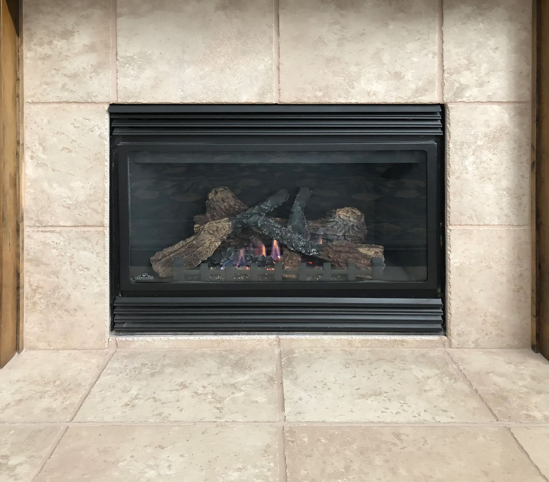 Gas fireplace with black frame, tan tile surround, and artificial logs inside.