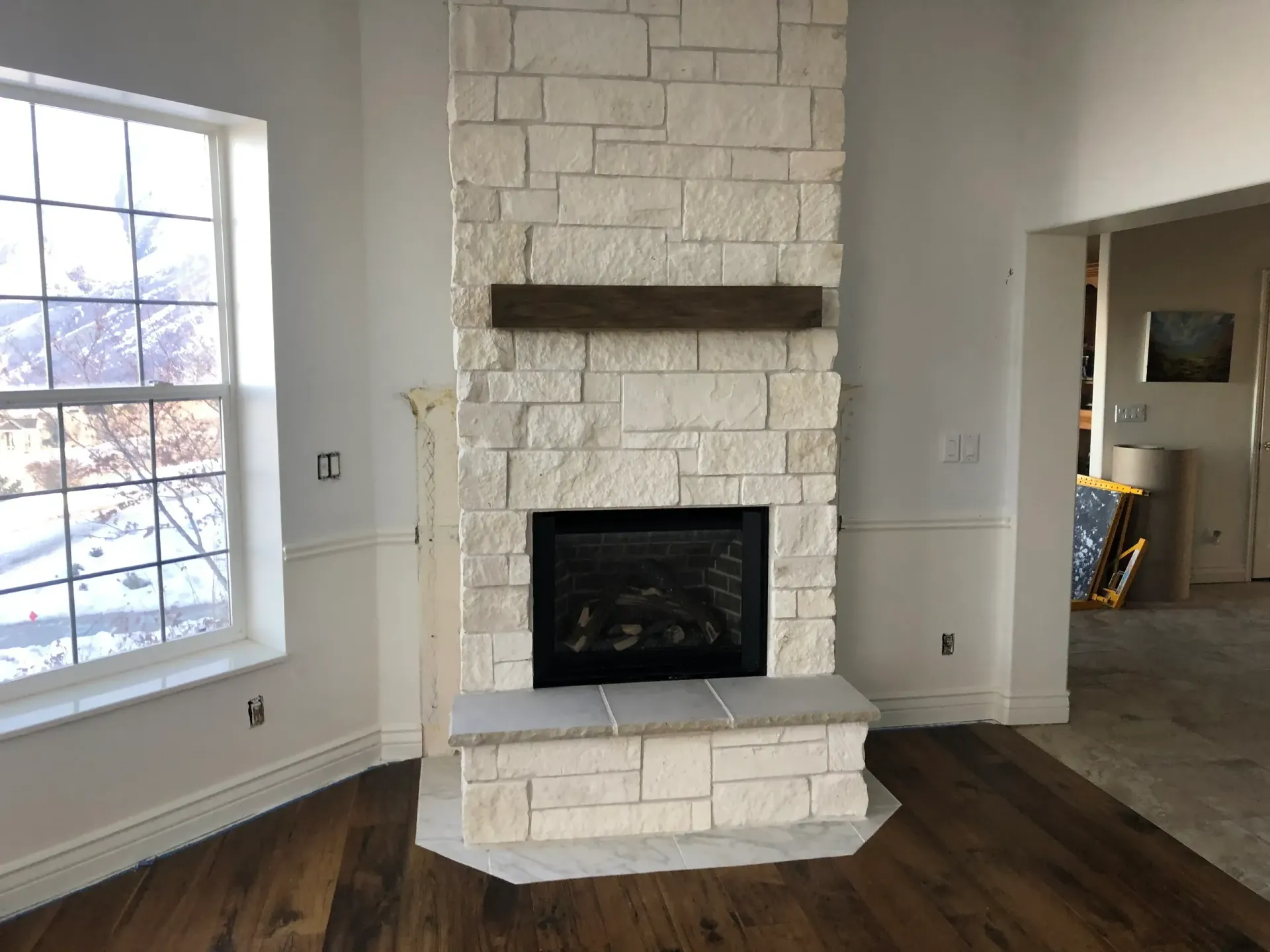 A fireplace with white stone and a wooden mantle in a white room with a large window and wood flooring.