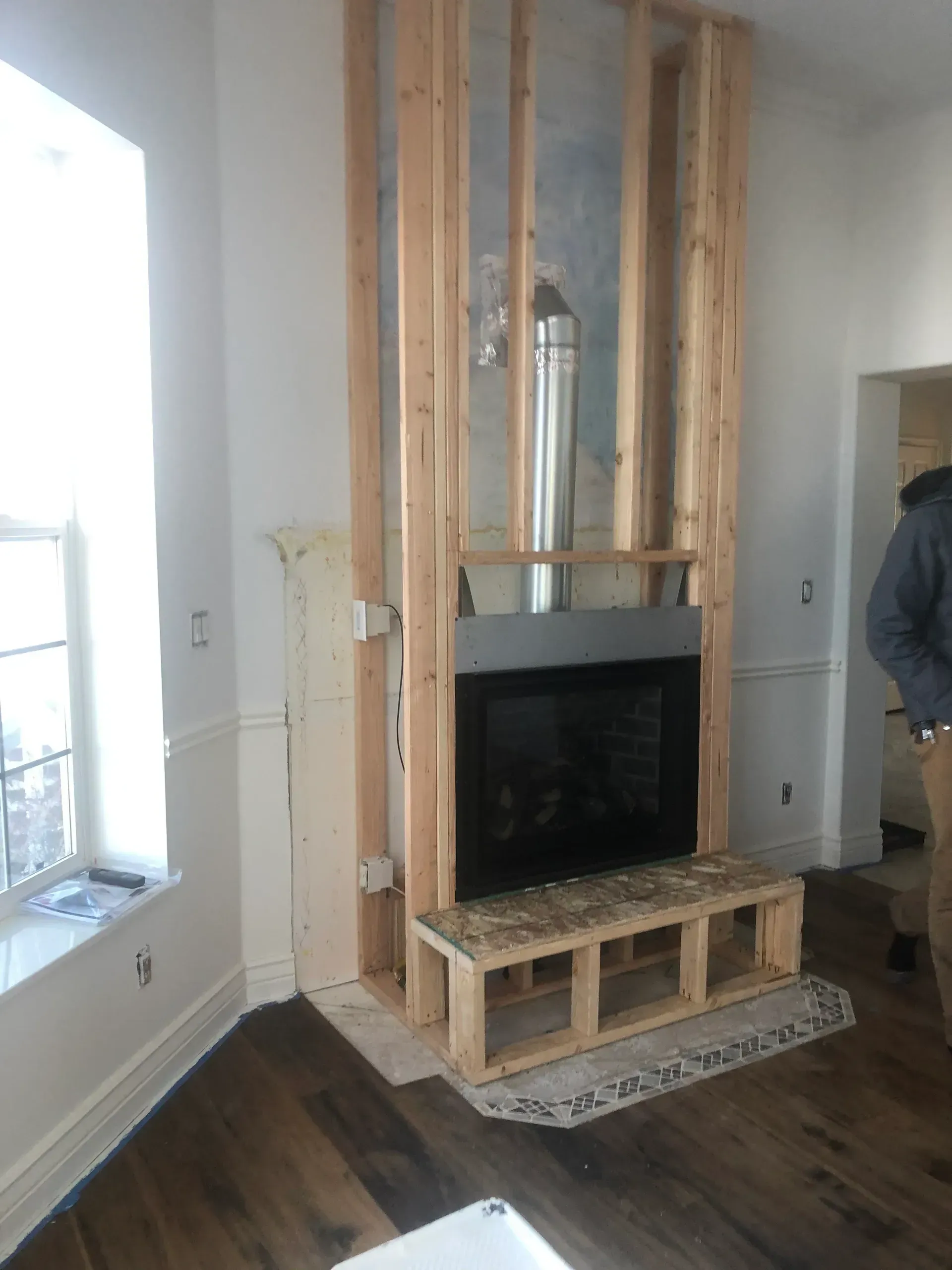 Fireplace under construction, wood frame, black insert, chimney.  Man standing in room with white walls, wood floors.