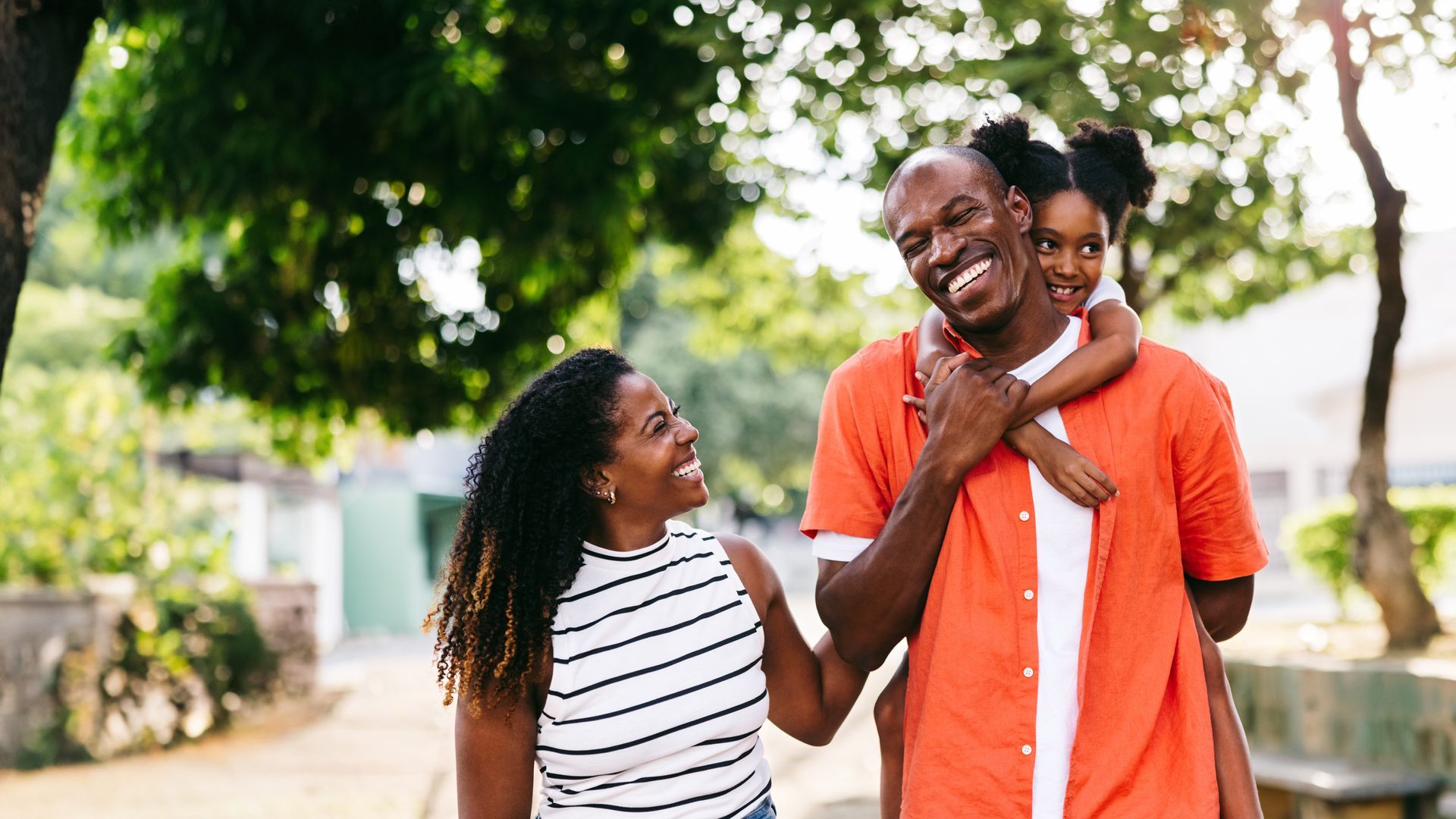 Family of three, smiling, walking outside. Father carries child on his back, mother walks alongside.