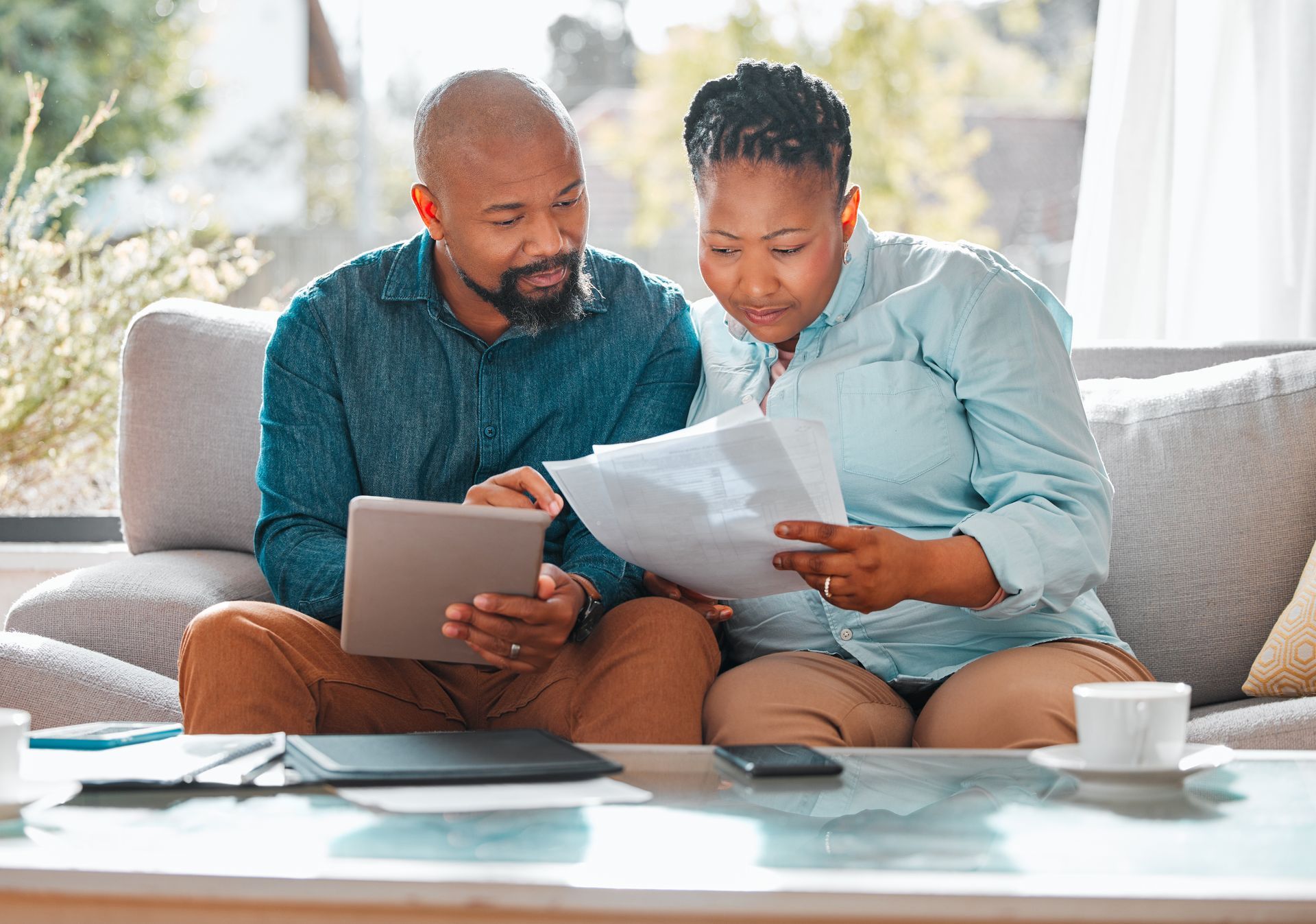 Couple on a sofa reviewing documents, man using a tablet, studying finances together.