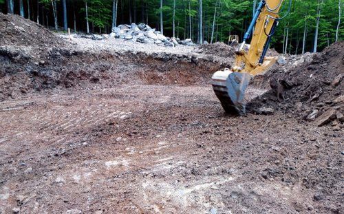 An excavator digs in a construction site with dirt mounds and trees in the background.