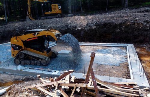 Yellow skid steer dumping gravel into a concrete foundation. Construction site, excavator in background.