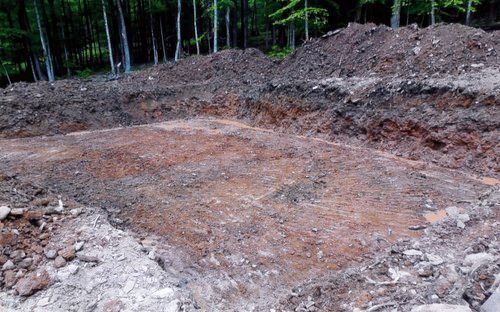 Excavated earth in a clearing, surrounded by piled soil and trees in the background.