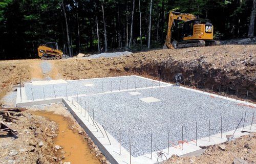 Construction site with concrete foundation, gravel fill, and heavy machinery; excavator in background.
