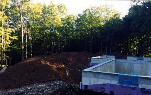 Construction site with concrete foundation and large dirt pile against a backdrop of trees.