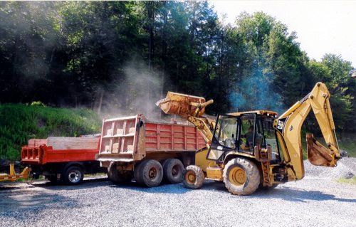 Yellow excavator loading red dump truck with gravel; outdoors, trees in background.