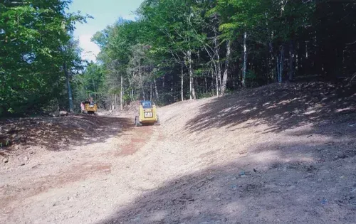 Two yellow skid steers on a dirt path in a wooded area, likely clearing land.