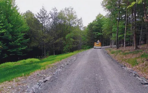 Gravel road through a forest with a yellow vehicle in the distance. Lush green trees line the sides.
